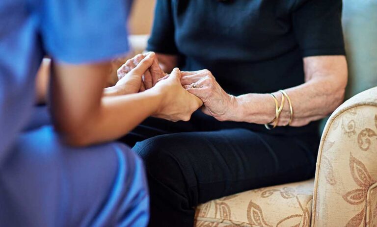 A caregiver reviews labeled medication cups and a resident’s medication chart at a dining table.
